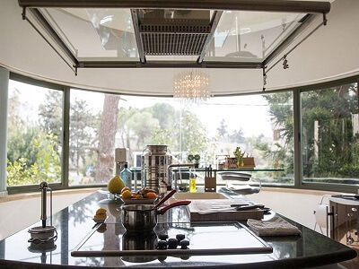 תמונה פנימית של בית מואר, שולחן מטבח עמוס בכלי מטבח ובאוכל, תקרה עם מנורהת חלונות שקופים  עם נוף לגינה